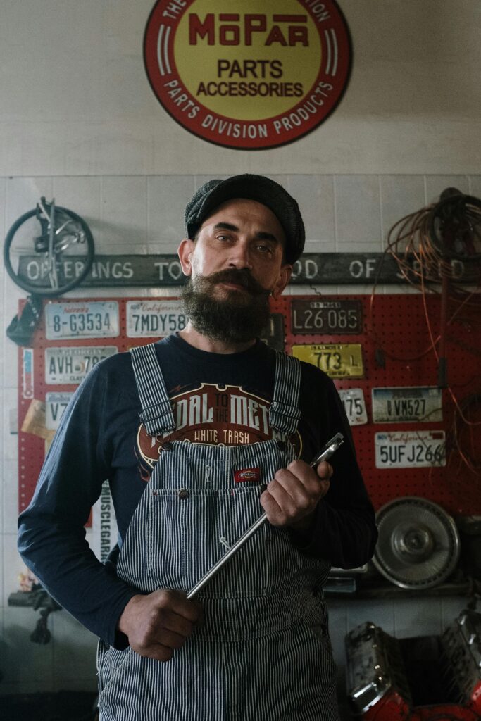 Bearded mechanic in overalls holding wrench in a garage adorned with license plates and vintage decor.